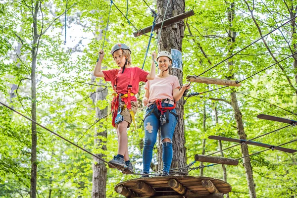 Mother and son climbing in extreme road trolley zipline in forest on carabiner safety link on tree to tree top rope adventure park. Family weekend children kids activities concept.