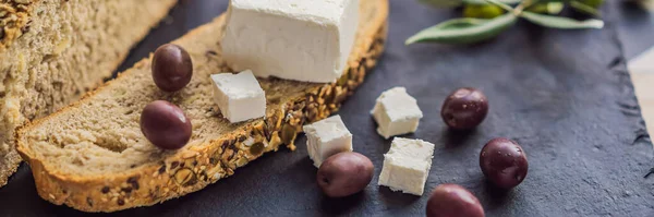 BANNER, LONG FORMAT Green and black olives with loaf of fresh bread, feta cheese and young olives branch on olive wood chopping board over dark background.