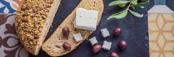 BANNER, LONG FORMAT Green and black olives with loaf of fresh bread, feta cheese and young olives branch on olive wood chopping board over dark background.