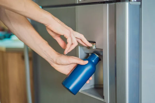 Male hand is pouring cold water and ice cubes in a metal bottle from dispenser of home fridge.