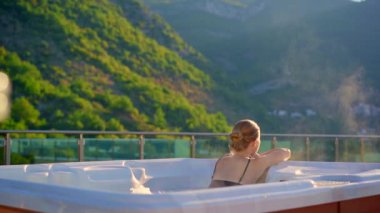 Yavaş çekim videosu. Genç bir kadın sıcak küvette dinleniyor. Çatıda, dağların manzarası var..