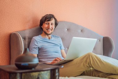 Adam yatakta oturuyor ve dizüstü bilgisayarla çalışıyor. Yatağın yanında Aroma ayırıcı.