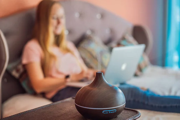 A woman sits on a bed and works on a laptop. Aroma diffuser next to the bed.