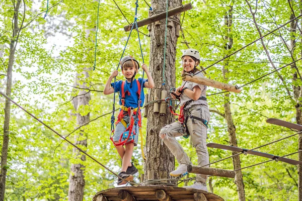 Mother and son climbing in extreme road trolley zipline in forest on carabiner safety link on tree to tree top rope adventure park. Family weekend children kids activities concept.