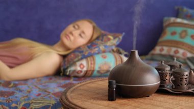 A young woman enjoys the atmosphere created by an aroma diffusor. She is laying on a couch with the lamp in the foreground. Aromatherapy Concept. Electric Ultrasonic Essential Oil Aroma Diffuser and