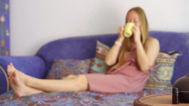 Genç bir kadın aroma difüzörünün yarattığı atmosferden zevk alır. Bir kanepede oturuyor. Önünde lamba olan bir bardaktan içiyor. Aromaterapi Konsepti. Elektrik Ultrasonik Temel Yağ