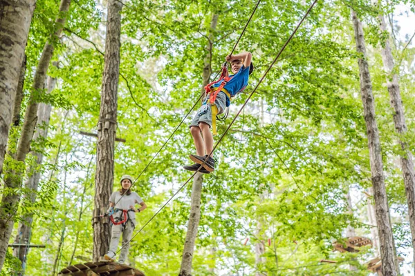 Mother and son climbing in extreme road trolley zipline in forest on carabiner safety link on tree to tree top rope adventure park. Family weekend children kids activities concept.