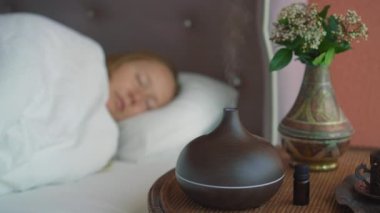 Genç bir kadın yatağında uykuya dalıyor ve aroma yayıcısının yarattığı atmosferin tadını çıkarıyor. Aromaterapi Konsepti. Elektronik Ultrasonik Temel Yağ Aroma Yayıcı ve Nemlendirici. Odaklanma değişiklikleri