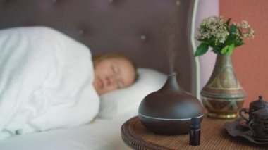 Genç bir kadın yatağında uyuyarak koku yayıcısının yarattığı atmosferin tadını çıkarıyor. Aromaterapi Konsepti. Elektronik Ultrasonik Temel Yağ Aroma Yayıcı ve Nemlendirici. Lambadan uzaklaşmaya odaklan
