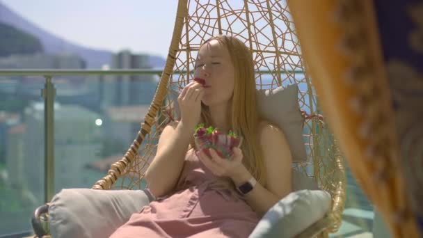 Een Jonge Vrouw Eet Aardbeien Zittend Een Schommelstoel Een Balkon — Stockvideo