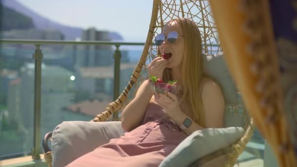 一位年轻女子正坐在以高山为背景的阳台上的摇椅上吃草莓 — 图库视频影像