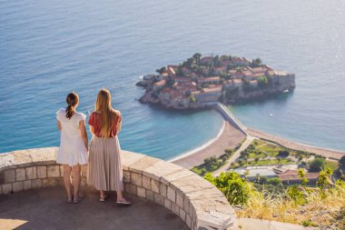 Two Woman tourist on background of beautiful view of the island of St. Stephen, Sveti Stefan on the Budva Riviera, Budva, Montenegro. Travel to Montenegro concept.