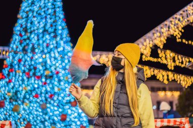 Pamuk şeker tutan ve Noel panayırında gülümseyen sarı yün şapka takan genç bir kadın Coronavirus COVID 19 'a karşı tıbbi maske takıyor..