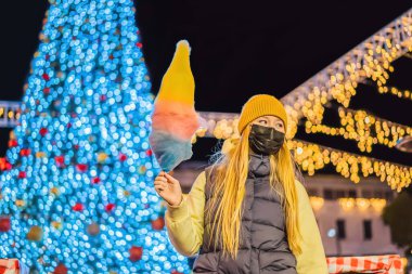 Pamuk şeker tutan ve Noel panayırında gülümseyen sarı yün şapka takan genç bir kadın Coronavirus COVID 19 'a karşı tıbbi maske takıyor..