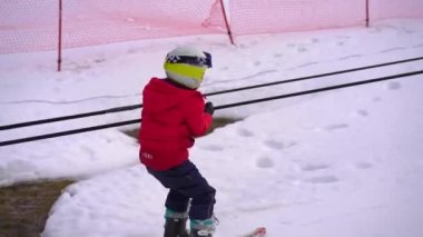Kırmızı ceket giyen küçük bir çocuk kayak yapmayı öğreniyor. Kayak asansörüyle tepeye çıkıyor ve yamaçtan aşağı iniyor..