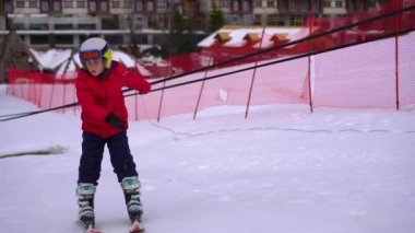 Kırmızı ceket giyen küçük bir çocuk kayak yapmayı öğreniyor. Kayak asansörüyle tepeye çıkıyor ve yamaçtan aşağı iniyor..