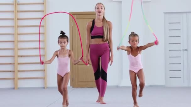 Две маленькие девочки гимнастки идут вперед, перепрыгивая через веревку, а их тренер наблюдает и следует за ними — стоковое видео