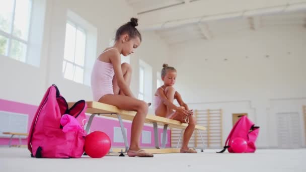 Entrenamiento de gimnasia - niñas sentadas en un banco en un pabellón deportivo y poniéndose zapatos de ballet — Vídeo de stock