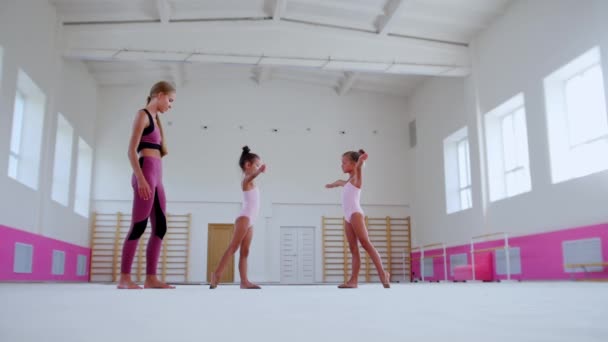 Jimnastik eğitimi - uzun boylu ve güzel bayan antrenör ve iki küçük kız öğrencisi büyük spor salonunda antrenman yapıyor. — Stok video