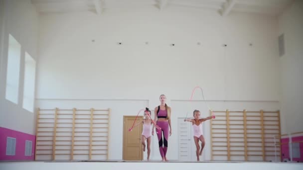 Spor salonunda jimnastik eğitimi. İpin üzerinden atlarken yürüyen iki küçük akrobatik kız ve onları izleyen ve izleyen kadın antrenörleri. — Stok video