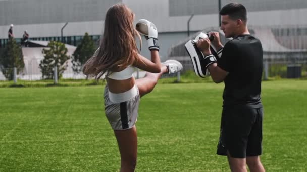 Young woman in boxer gloves having a training outdoors and kicking her trainer in the mitt — Stock Video