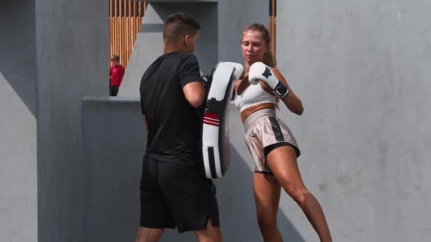 Молодая женщина в белом топе, занимающаяся боксом со своим тренером на улице - пинание портативной боксерской груши — стоковое видео
