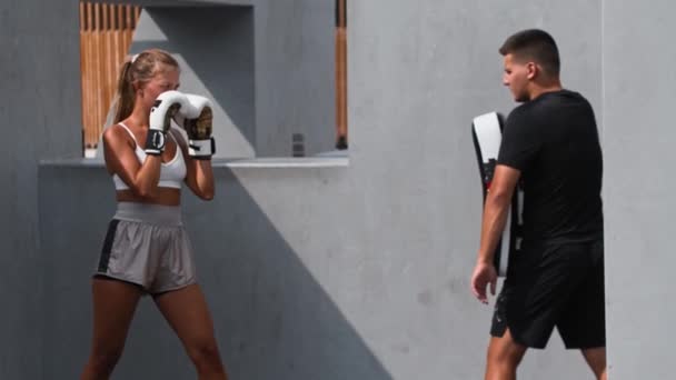 Koçuyla boks antrenmanı yapan genç bir kadın portatif kum torbasını tekmeliyor. — Stok video