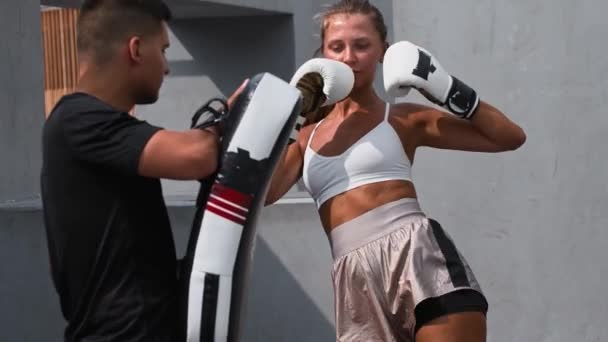 Молода жінка в білому верхній частині має тренування боксу зі своїм тренером зовні - штовхаючи портретний мішок з повороту — стокове відео