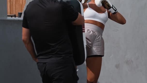 Beyaz elbiseli genç bir kadın dışarıda boks antrenmanı yapıyor. Koçuna bağlı duran kum torbasını tekmeliyor. — Stok video