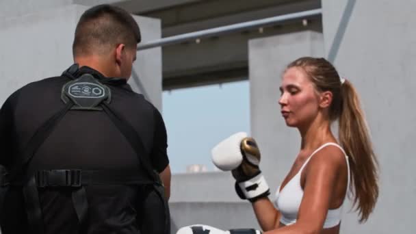 Young woman in white top having a boxing training outdoors with a male coach - punching the protectors on her coachs body — Stock Video