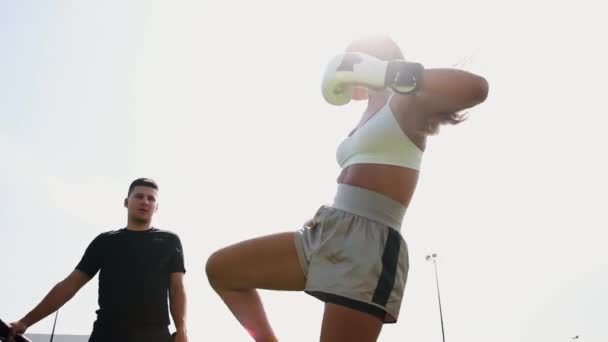 Entrenamiento de boxeo al aire libre - mujer joven pateando el objetivo en su mano de entrenadores — Vídeos de Stock
