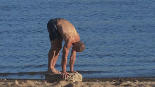 Los ejercicios deportivos al aire libre - el hombre joven en forma comienza a pararse en sus manos sobre la piedra en una playa — Vídeos de Stock