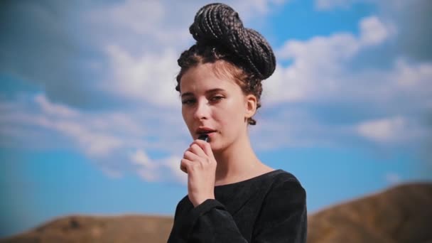 Young beautiful woman with dreadlocks standing on mountain - vaping and exhales steam - looking in the camera — Stock Video