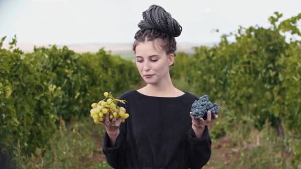 Młoda kobieta z dredami trzymająca białe i czarne winogrona w dłoniach i patrząca w kamerę — Wideo stockowe