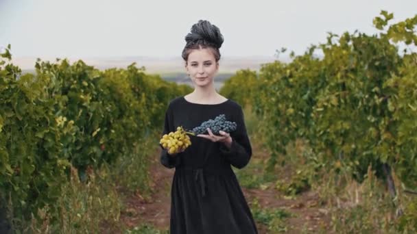 Jovem mulher sorridente com dreadlocks segurando uvas brancas e pretas em suas mãos e olha para a câmera — Vídeo de Stock