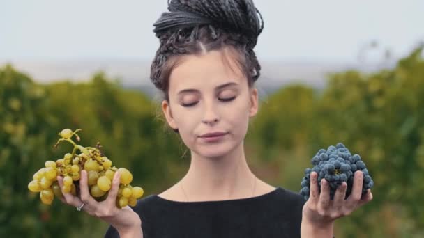 Jovem com dreadlocks segurando uvas brancas e pretas em suas mãos e morde alguns pedaços — Vídeo de Stock