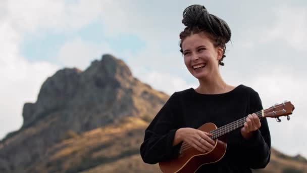 Mladá smějící se žena s dredy přivázanými k hlavě hraje ukulele v horách