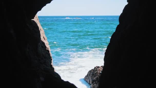 Zicht vanuit de donkere grot op de blauwe zee en golven die neerstorten op rotsen — Stockvideo