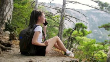 Rastalı genç bir kadın yürüyüşçü ormanda yerde oturuyor ve telefonda tatilleri hakkında konuşuyor.