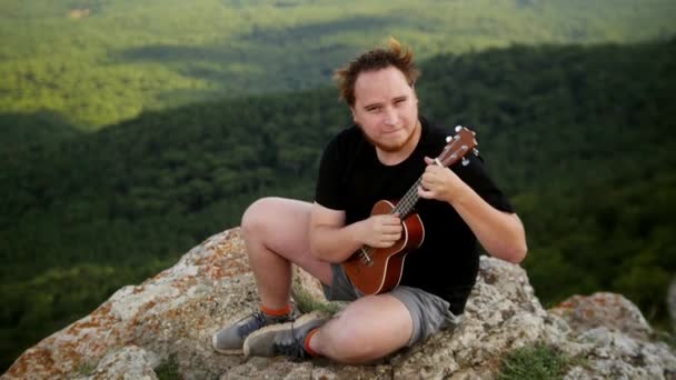 Przygoda - młody rudy człowiek turysta siedzi na szczycie góry i gra ukulele — Wideo stockowe