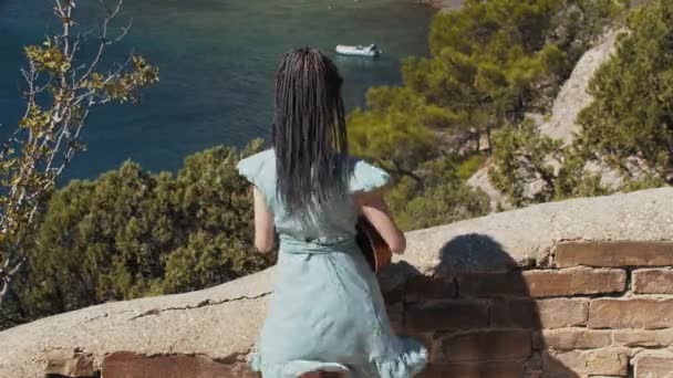 Une jeune femme avec dreadlocks debout près du mur de briques avec une vue sur la baie de la mer et de jouer ukulélé — Video