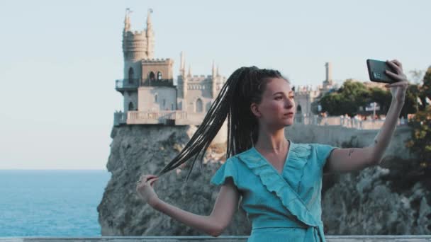 Молодая женщина с дредами, стоящая на фоне моря и замка на краю скалы - фотографирует себя — стоковое видео