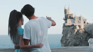 Genç bir çift denizin arka planında dikiliyor ve uçurumun kenarında bir kale ve bunu tartışıyor.