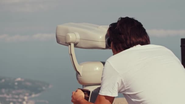 Young man looks through a telescope from a viewpoint on a sea vacation — Stock Video