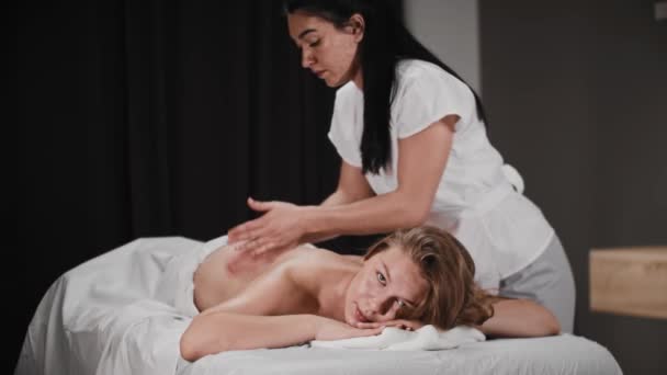 Mladá žena dostává masáž od ženské masérky s černými vlasy a dívá se do kamery — Stock video