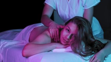 Neon ışıklı masaj seansı. Kanepede uzanmış masaj yaptıran ve kameraya bakan genç bir kadın.