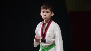 Beyaz üniformalı küçük bir çocuk tekvando yapıyor. Pozisyon alıyor ve hareketlerini gösteriyor.