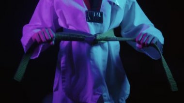 Neon ışıkları altında tekvando yapan küçük bir çocuk yeşil kuşağını bağlıyor ve elini sallıyor.