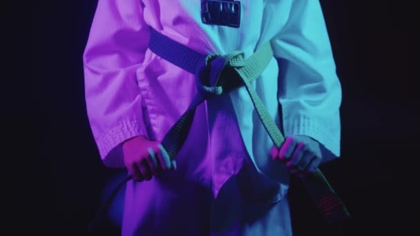 En liten pojke gör taekwondo i neon belysning - binda upp sitt gröna bälte på midjan — Stockvideo