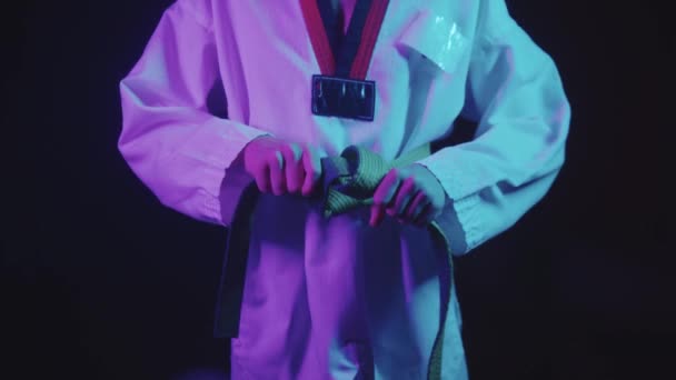 彼の緑のベルトを結ぶとカメラを見て-ネオンの照明でtaekwondoをやっている小さな男の子 — ストック動画